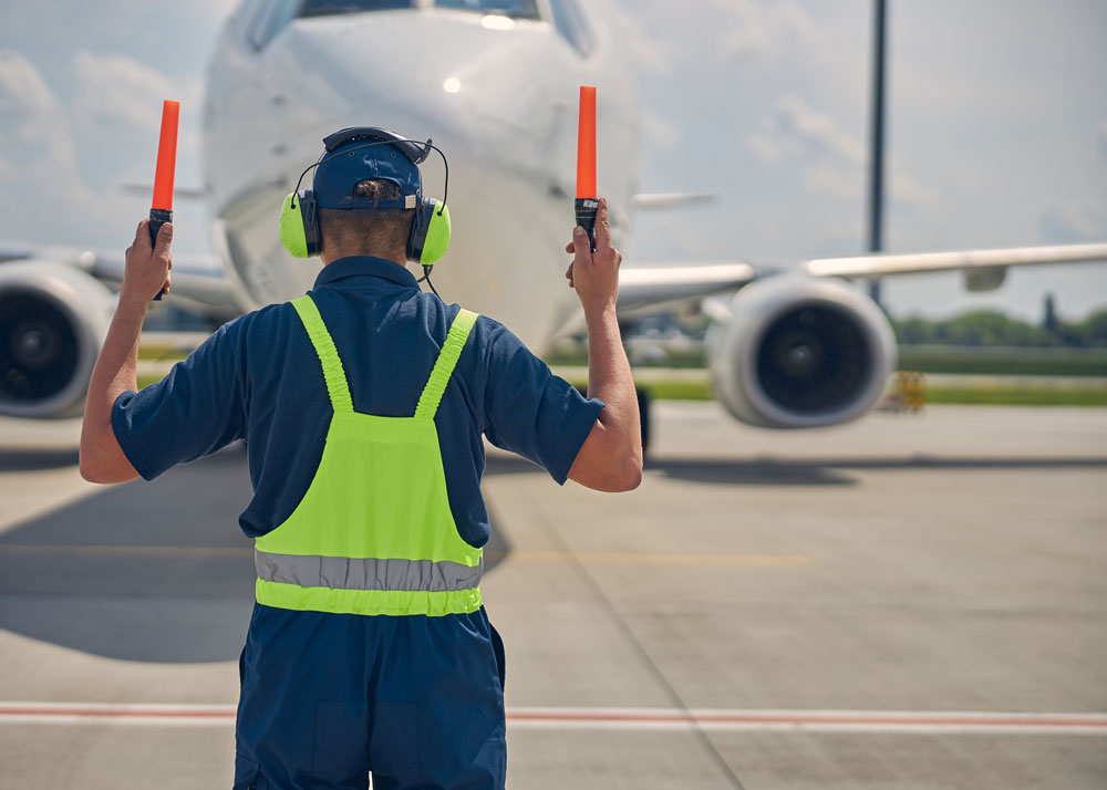 An airplane marshall wearing blue coveralls and bright green reflective vest directs an airplane onto the tarmac with orange directional wands.