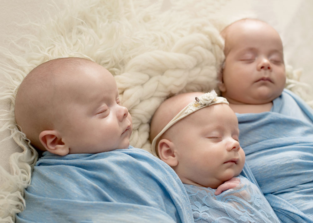 three babies, triplets, wearing blue pajamas sleep on a white blanket.