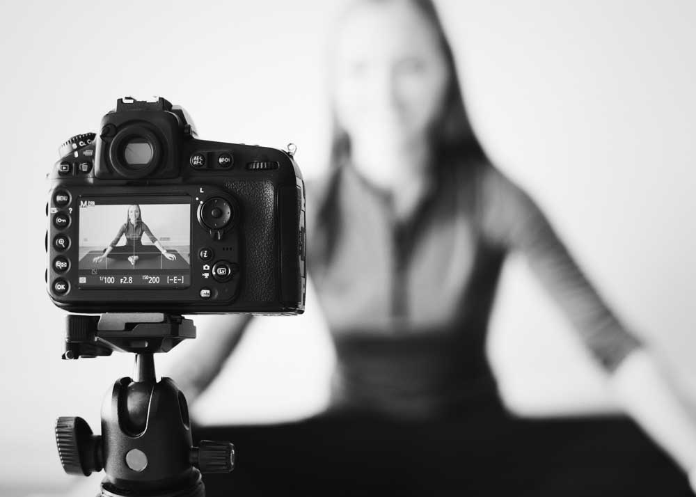 A black and white photo of a woman doing a video recording of herself. The image of the woman is clear in the camera's viewfinder, but blurred in the background.